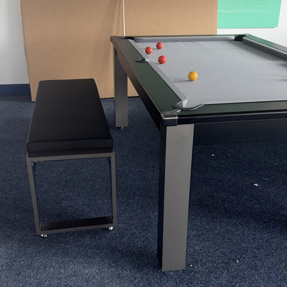 A modern pool dining table with a gray felt surface, black frame, and matching black cushioned bench, set on a blue carpeted floor in an indoor room.
