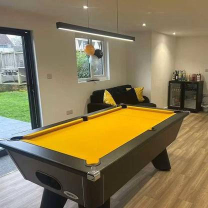 A yellow felt pool table in a modern home game room with wood-look flooring, a black sofa with yellow pillows, and a small bar cart in the background.